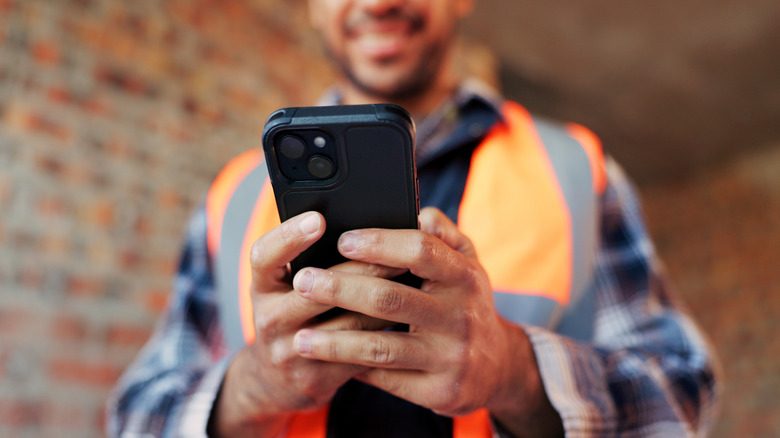 Photo from an upward angle of a man holding a black smartphone. His hands are at the center, while the rest of his figure and the brick walls are out of focus.