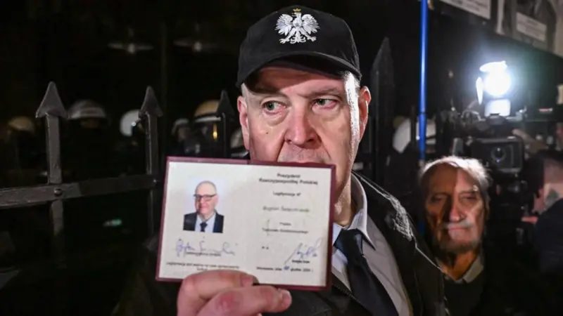 Bogdan Święczkowski sostiene una identificación con su foto frente a una valla metálica. Lleva una gorra de béisbol negra con el símbolo de un águila blanca. En la imagen nocturna se aprecian otras pe