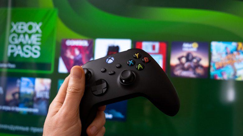Xbox controller held up in front of a screen displaying Xbox Series X home page.