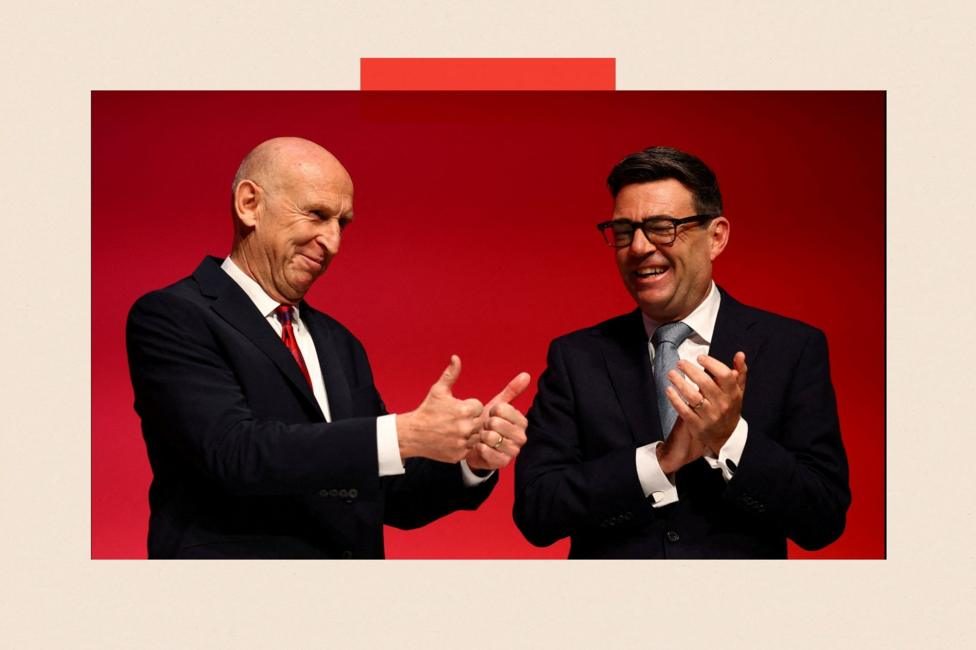 John Healey and Andy Burnham. Healey is doing a thumbs up with both hands and Burnham is clapping. They are wearing suits and are standing in front of a red background.