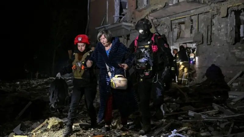 Plano general que muestra a una mujer de mediana edad alejándose de un edificio residencial dañado por ataques rusos durante la noche. Va de la mano de dos rescatistas que la ayudan a caminar sobre lo