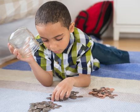 A boy lying on the floor counts coins from a glass jar on a rug