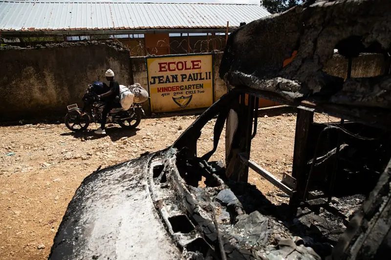 Un hombre monta una motocicleta pasando por los restos de un vehículo carbonizado después de la violencia nocturna en la que más de veinte personas fueron asesinadas por bandas armadas en Kenscoff, un